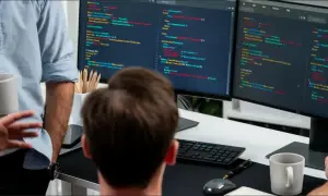 Dwóch programistów pracuje nad kodem. Jeden trzyma kubek, drugi wskazuje na ekran z kodem. Widoczny jest zaawansowany stos technologiczny.