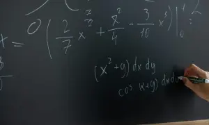 Rysunek przedstawia tablicę z równaniami matematycznymi. Widać całki i wzory, a ręka pisze markerem.