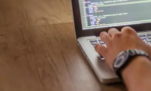 Dłoń pisze kod w języku php na laptopie. Widać fragmenty kodu CSS i ścieżkę do pliku.