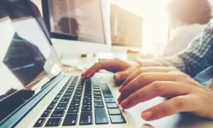 Osoba pisze na laptopie, ucząc się, jak zaindeksować stronę w Google. W tle monitory z kodem.