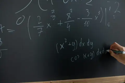 Rysunek przedstawia tablicę z równaniami matematycznymi. Widać całki i wzory, a ręka pisze markerem.