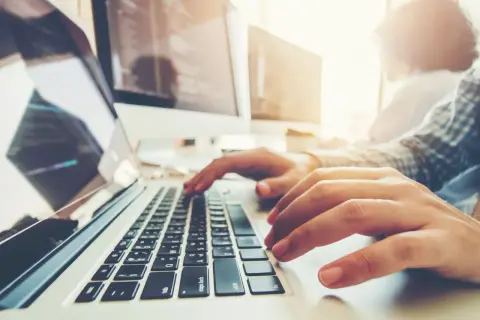 Osoba pisze na laptopie, ucząc się, jak zaindeksować stronę w Google. W tle monitory z kodem.