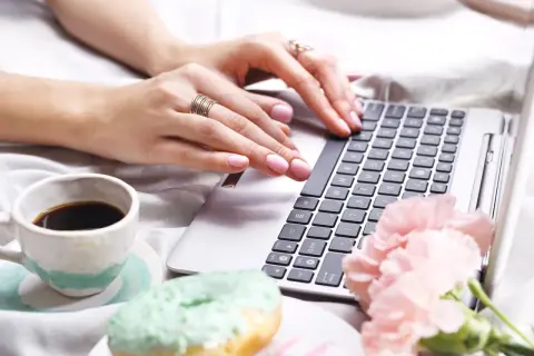 Kobieta pisze na laptopie, obok kawa i pączki. Dowiedz się, jak usunąć twardą spację, by tekst wyglądał idealnie.