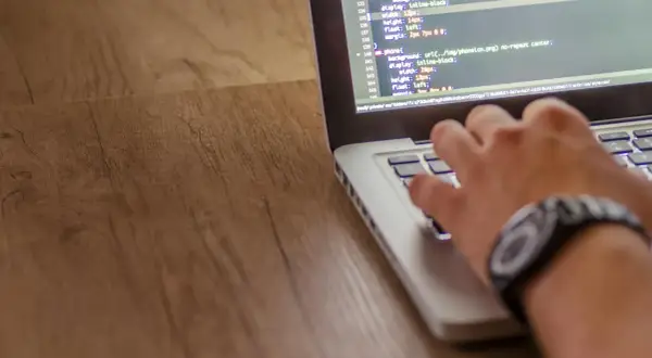 Dłoń pisze kod w języku php na laptopie. Widać fragmenty kodu CSS i ścieżkę do pliku.