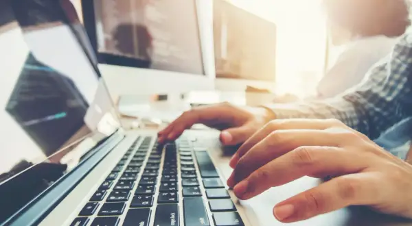 Osoba pisze na laptopie, ucząc się, jak zaindeksować stronę w Google. W tle monitory z kodem.