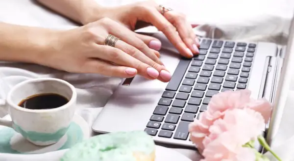 Kobieta pisze na laptopie, obok kawa i pączki. Dowiedz się, jak usunąć twardą spację, by tekst wyglądał idealnie.