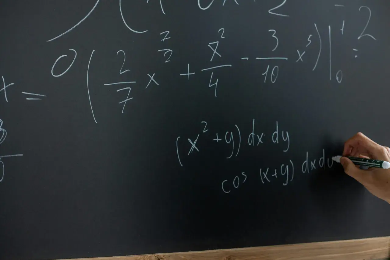 Rysunek przedstawia tablicę z równaniami matematycznymi. Widać całki i wzory, a ręka pisze markerem.