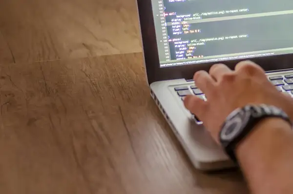 Dłoń pisze kod w języku php na laptopie. Widać fragmenty kodu CSS i ścieżkę do pliku.