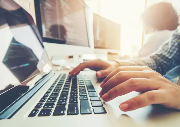 Osoba pisze na laptopie, ucząc się, jak zaindeksować stronę w Google. W tle monitory z kodem.