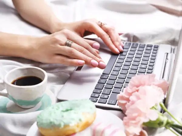 Kobieta pisze na laptopie, obok kawa i pączki. Dowiedz się, jak usunąć twardą spację, by tekst wyglądał idealnie.